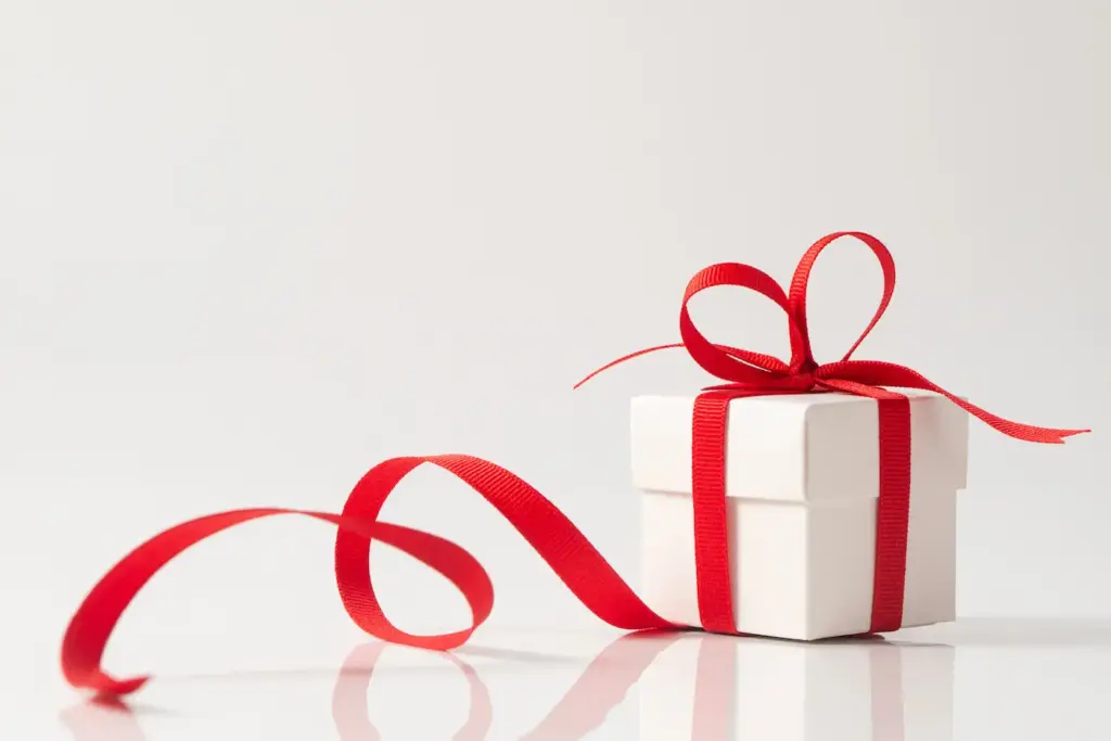 A small square white gift box tied with a red satin ribbon that forms a perfect bow on top, for Christmas, New Year or birthday theme. The ribbon trails around the box on a reflective white surface.