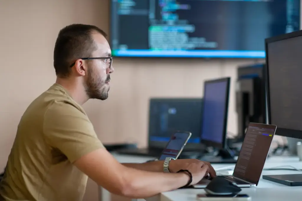 Softverski inženjer u uredu radi na prijenosnom računalu dok se u pozadini nalaze dodatni monitori s kodom i prikazom sustava
