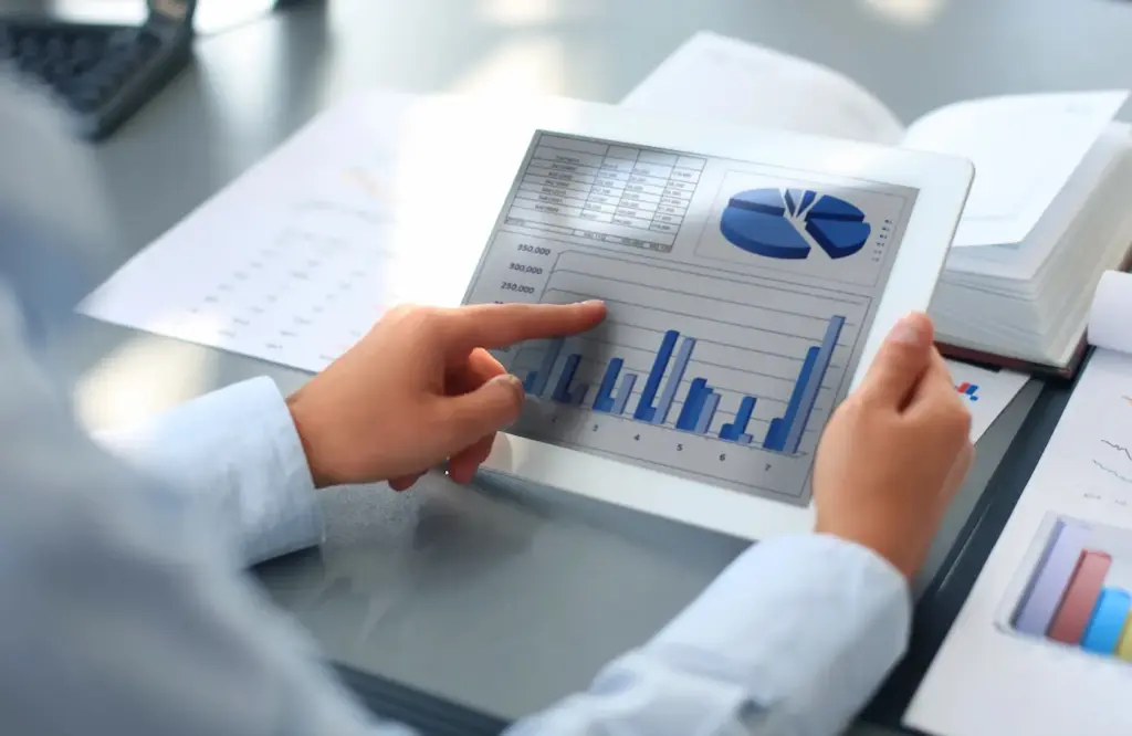 Krupni plan ruku koje drže tablet s prikazom poslovnih statistika i grafikona dok u pozadini stoje otvoreni registratori i financijska izvješća